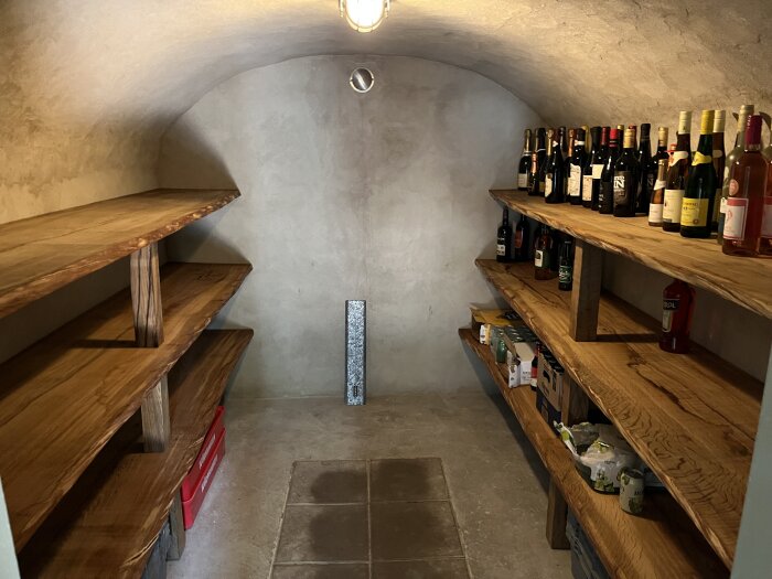 Jordkällare med trähyllor fyllda med flaskor och diverse varor. Golvet är delvis i cementplattor. Lampan i taket lyser upp rummet.