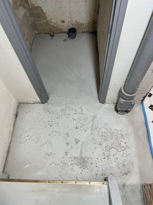 Nyflytspacklat golv i litet rum med två dörröppningar, synliga väggskador och rör i ena hörnet.