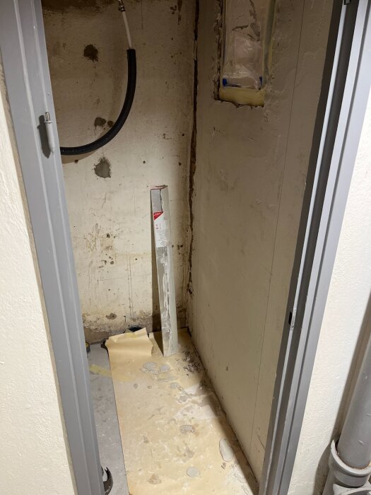 Litet utrymme under renovering med gipsade väggar och förberett för eldragning, plastskyddat golv och rör längs vägg. En ventilationskanal syns.