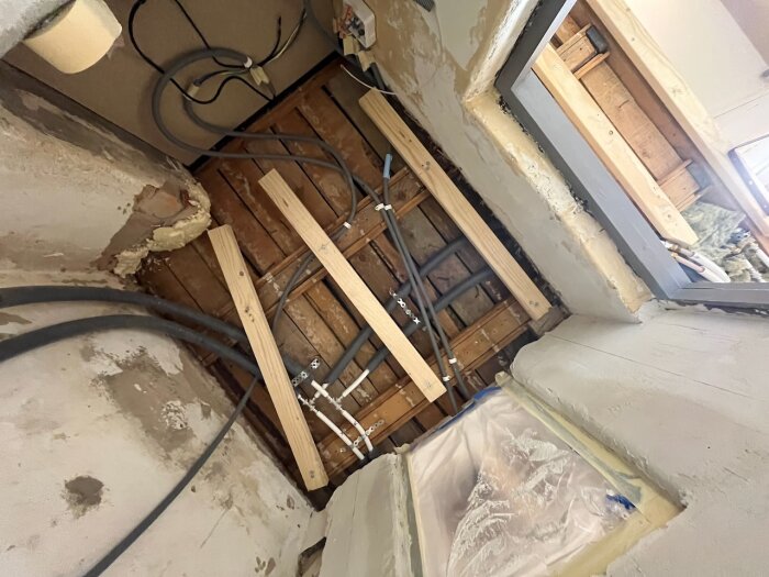 Rum under renovering med synliga träreglar och rördragning i taket. Reparerade väggar med gips och plastfodrad öppning mot yttervägg.