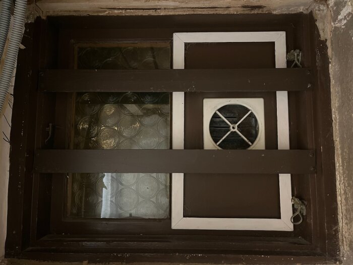 Ett gammalt fönster med två bruna träbrädor framför och en vit ventilationstrumma.