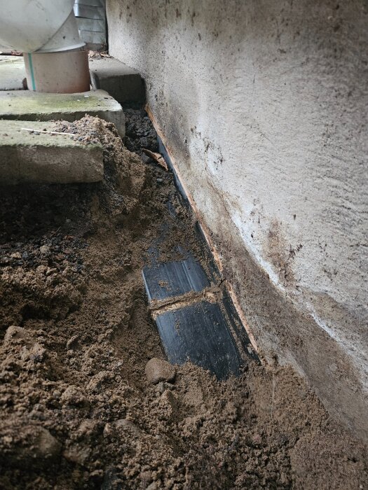 Jord som täcker en svart plaströrskarv vid en betongvägg. Eventuell orsaksplats för problem i ett byggprojekt.