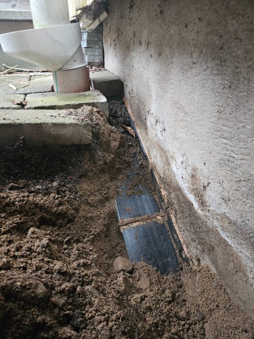 Skarv av dräneringsrör vid husgrund med synlig jord kring och potentiell läcka.