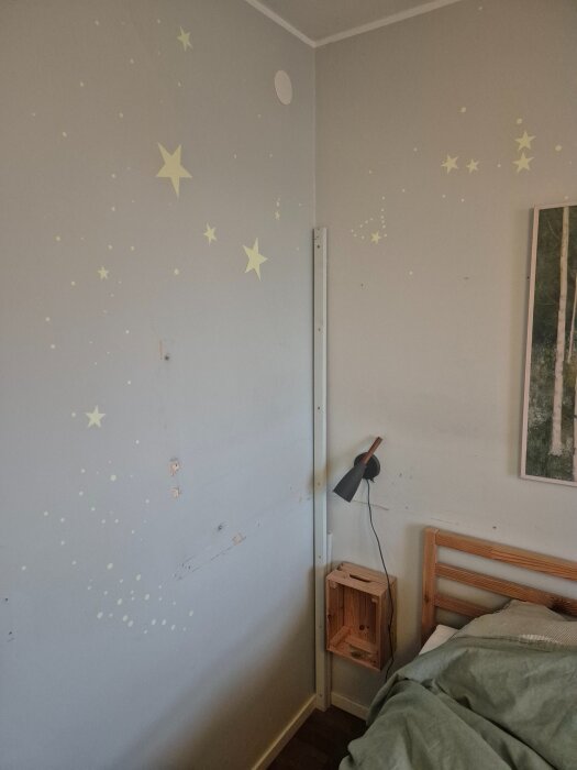 Vägg med lösa tapeter och stjärnor dekorerade, en svart vägglampa bredvid en säng, trädetaljer och ett träramshylla.