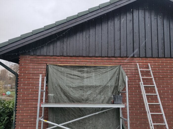 Ställning och stege mot en tegelvägg under svart träpanel, täckt fönster med presenning, byggprojekt pågår.