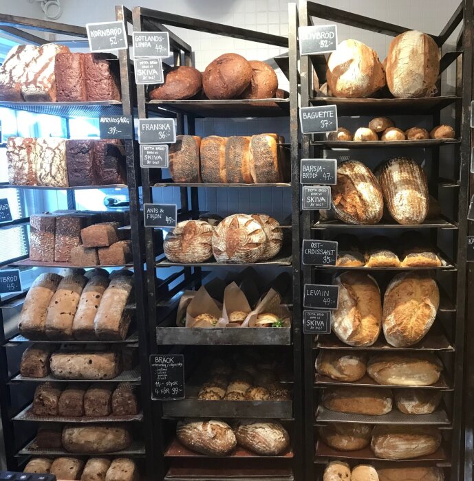 Brödbutik med olika sorters bröd på hyllor, inklusive levain, baguette och franska, med priser angivna på skyltar.