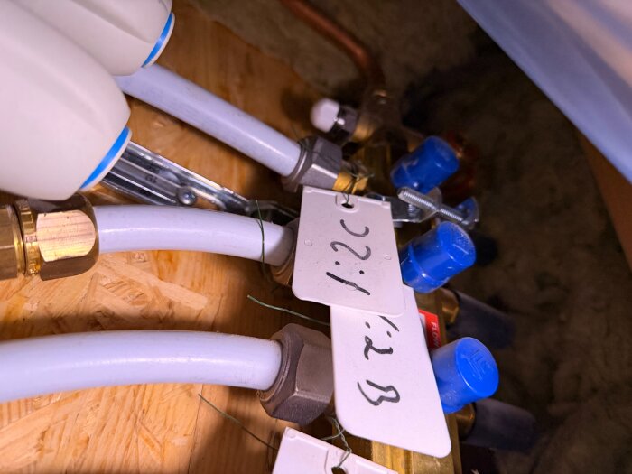 Golvvärmesystem med flera ledningar och blå ventiler på en träram, märkta med etiketter. Möjligen luftningsventiler synliga för service.