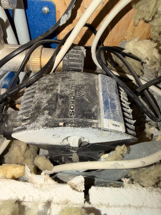 Cirkulationspump med rör och isolering i golvvärmesystemet, med dammig yta och omgivande kablar.