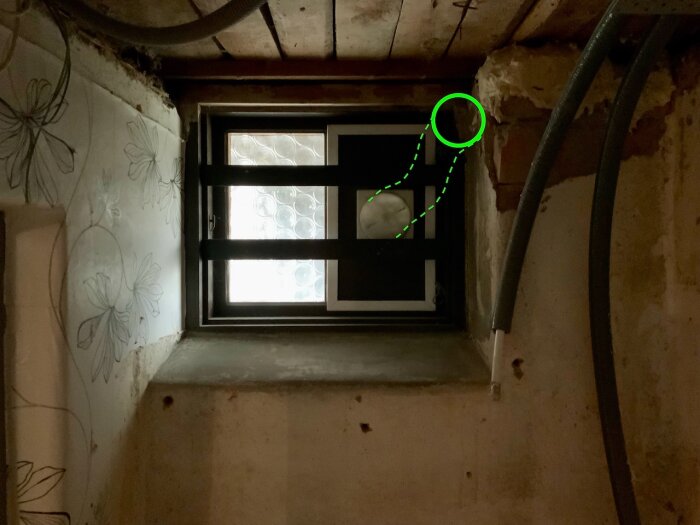 Gammalt fönster igenbyggt med svart bräda och nyinsatt ventil, omgiven av betongväggar med blommigt mönster och flexrör på sidan.