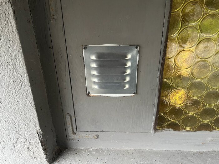 Monterad ventil på grå vägg bredvid glasparti med cirkulärt mönster.
