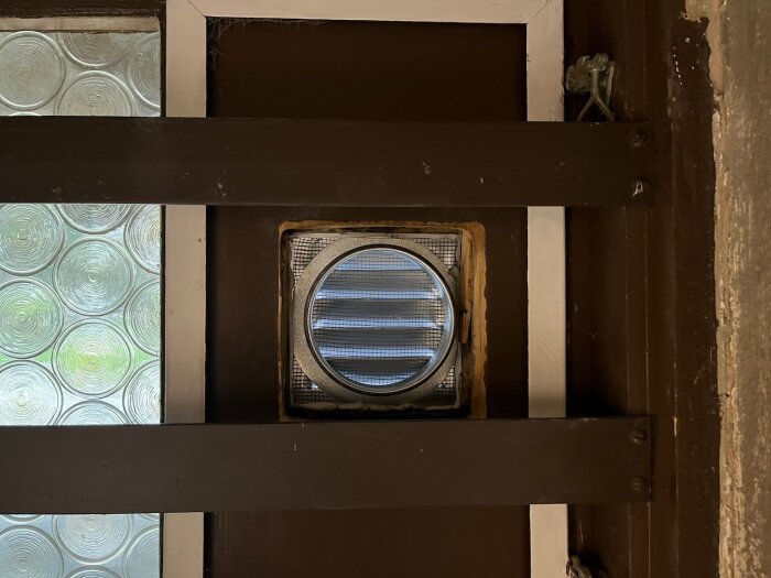 Ventil monterad på en mörk väggbredvid glasparti med cirkulära mönster. Ventilen omgiven av skruv och fogning, delvis dold bakom tvärgående brädor.