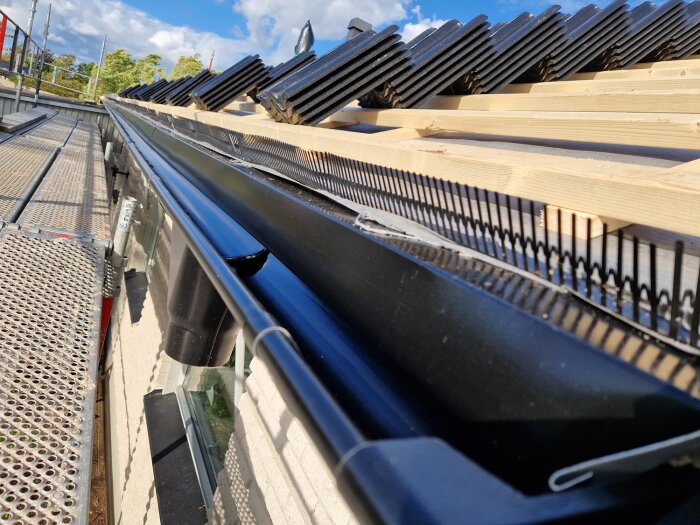 Nylagt tak med svarta takpannor och ny hängränna, fotograferat från sidan på en byggställning under klarblå himmel.