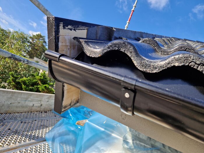 Svart tak med vågformade plattor och vattenränna på en blå himmel.