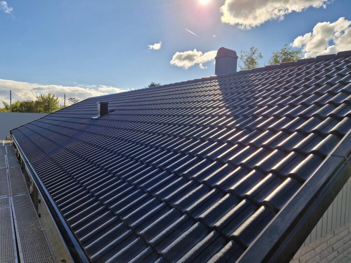 Svart tak med glänsande takpannor under molnig himmel och solljus.