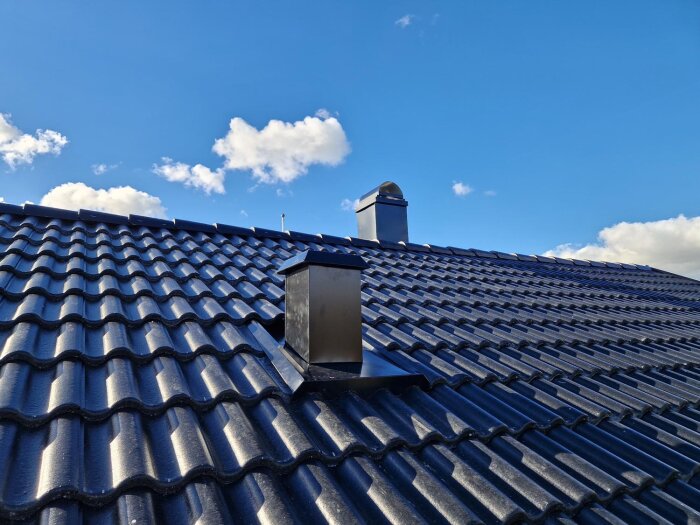 Svart tak med takpannor och två skorstenar under blå himmel med moln.