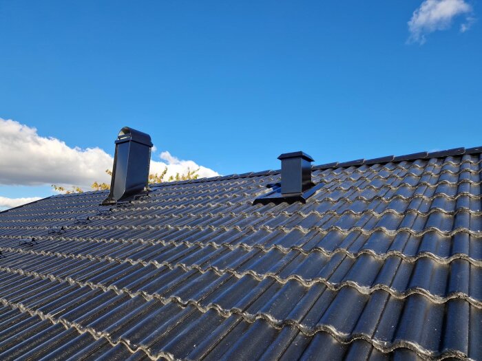 Tak med svarta takpannor och två ventilationsrör under blå himmel.