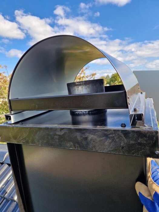 Svart ventilationshuv på tak med metallkupol under klarblå himmel, hand i blå handske vid kanten.