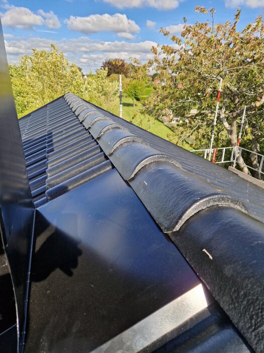 Svart tak med glaserade takpannor under klar blå himmel, omgiven av grönskande träd.