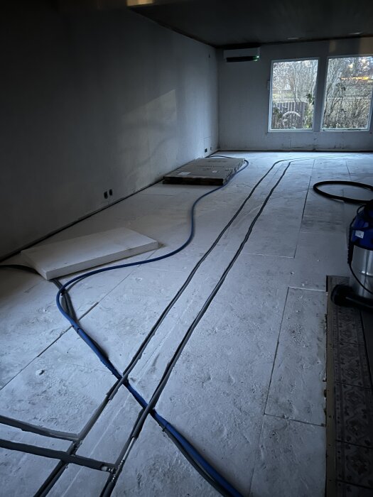 Rum under renovering med synliga golvkablar och styrofoamlåda, stort fönster i bakgrunden.