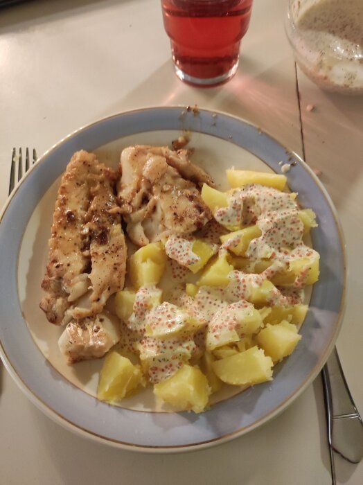 Smörstekt torsk med kokt potatis och hemgjord romsås på en tallrik, bredvid ett glas röd dryck.