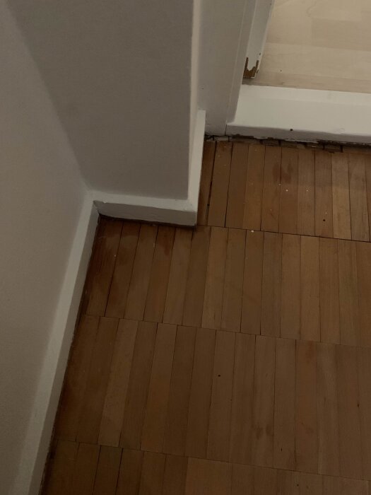 Parkettgolv i sovrum med smärre glipor och mörkare fläckar vid dörröppning; behov av renovering synligt.