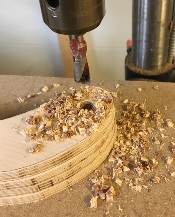 Borrmaskin borrar genom flera lager av träplywood, omgiven av träflis och spån på en arbetsbänk.