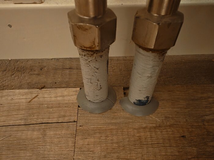 Två rör med muttrar upptill, placerade tätt intill varandra ovanpå ett trägolv. Rören ser slitna ut med oregelbundna ytor.