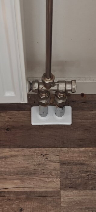 Värmeledning med metallrör och kopplingar, installerade mot en vägg och ett trägolv. Två rör syns med en gemensam täckbricka.