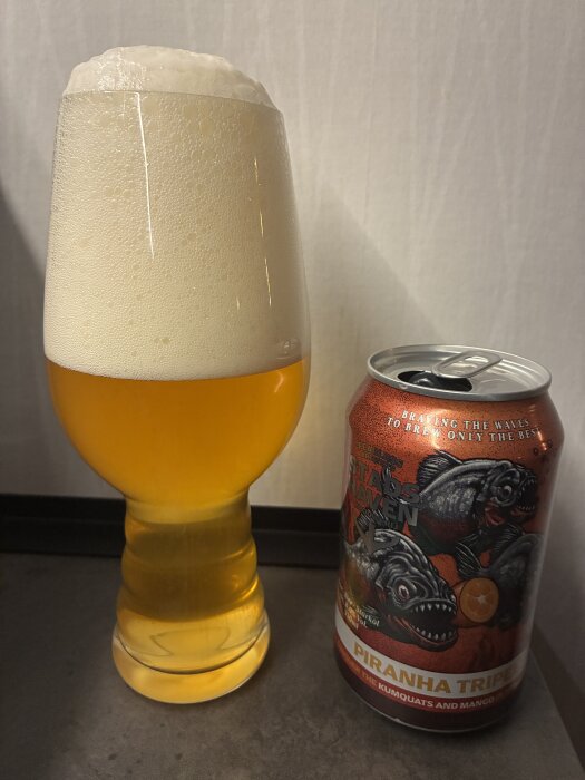 Ölglas med skumkrona bredvid en burk "Piranha Triple" öl, med etikett av pirayor, kumquats och mango.