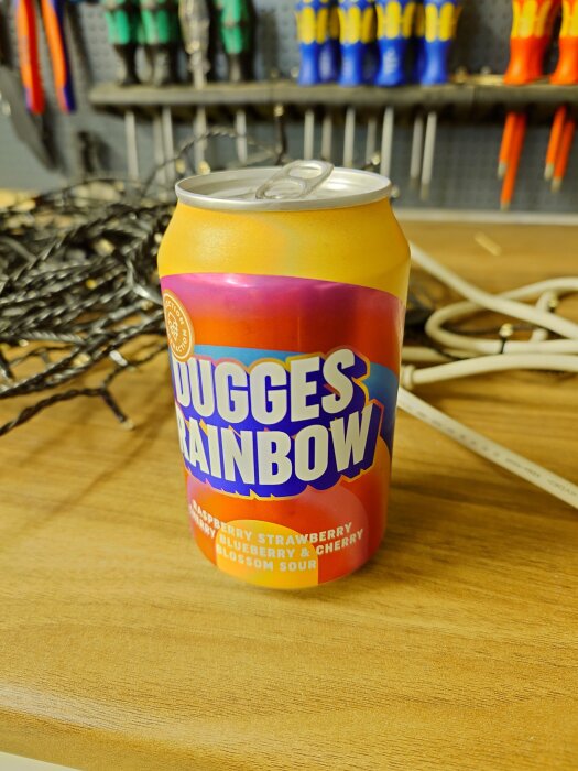 En färgglad läskburk med texten "Dugges Rainbow" står på ett träbord, omgiven av verktyg i en verkstadsmiljö.