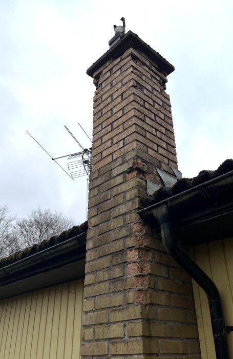Tegelstenskorsten med sprickor och antenn, delvis ersatta tegelstenar. Renovering planeras genom putsning för långsiktig hållbarhet.