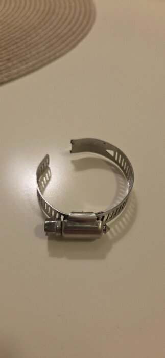 Avbildad är en trasig slangklämma av metall liggande på en ljus yta.