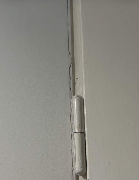 Målad dörrkarm med ojämnt spackel, luckor och synliga sprickor längs av dörrhängsletillhörande väggen.