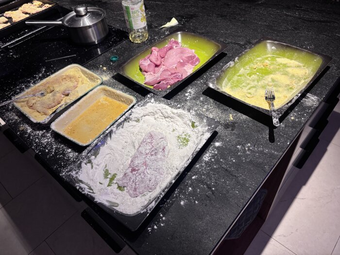 Tillagning av fläskschnitzlar på köksbänk med ingredienser som mjöl, ägg och rått kött i olika skålar, samt en kastrull på spisen.