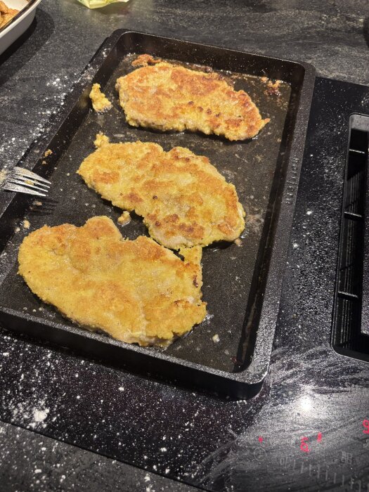 Tre gyllenbruna fläskschnitzlar ligger på en svart stekhäll. Det finns mjölrester runtomkring.