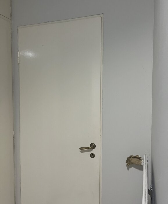 Vit dörr med guldfärgat handtag i hallen, omgiven av ojämnt målade väggar. Papper syns i dörrens närhet.