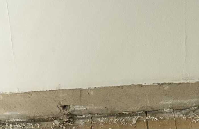 Betongvägg med "bucklor" och silvriga ringar är synliga, vägg nära golvet visar nötning och några utstickande delar som kan vara pluggar.