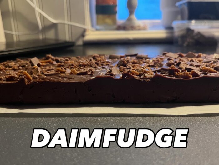 Hemgjord Daimfudge liggande på ett köksbord, toppad med hackad Daim och texten "DAIMFUDGE" över bilden.