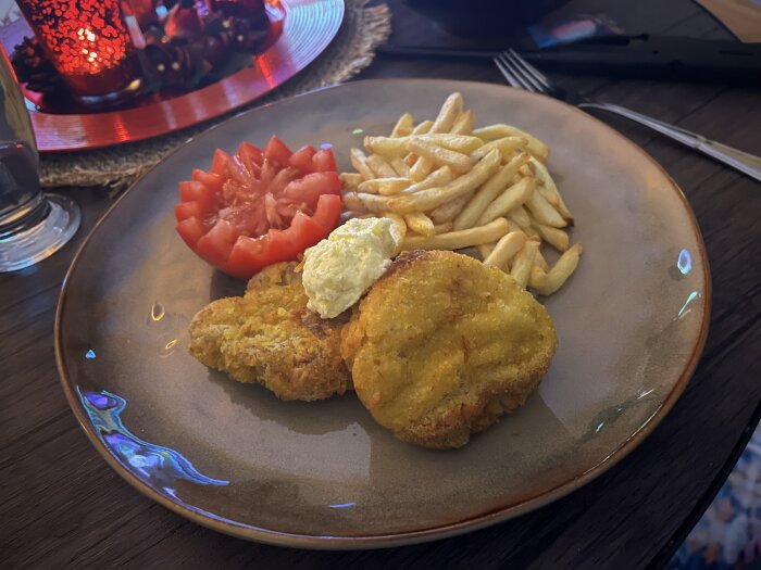 Tallrik med panerade biffar, pommes frites, tomatklyftor och en klick sås, placerad på ett bord med rött ljusdekoration i bakgrunden.