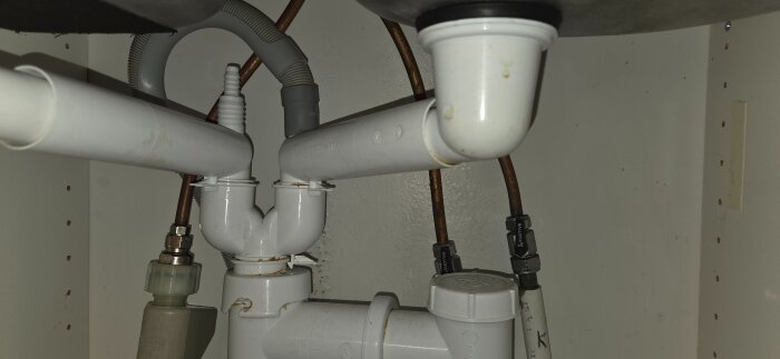 Vitt avloppsrörsystem under diskbänk med kopplingar och ventiler.