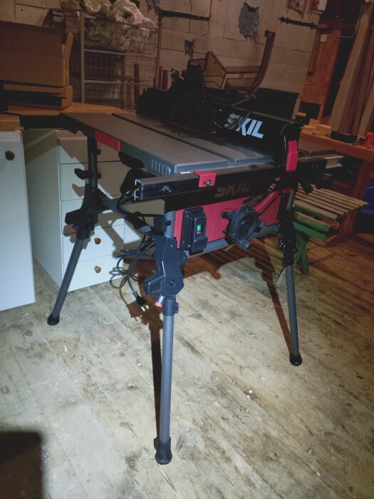 Bordssåg av märket Skil stående på golvet i en verkstad med träväggar och diverse byggmaterial i bakgrunden.