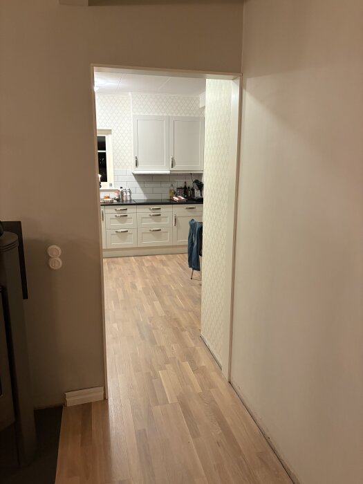 Kök med vita skåp och ljust trägolv, sett genom en dörröppning. Köksbänken har kakel ovanför och en stol syns delvis.