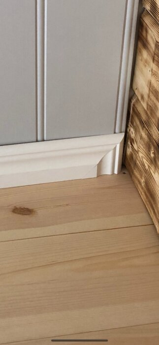 Vit golvlist vid grå vägg och träpanel.