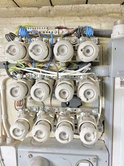 Bild på gammalt elskåp med flera porslinsäkringar och slingrande kablage mot en vägg.