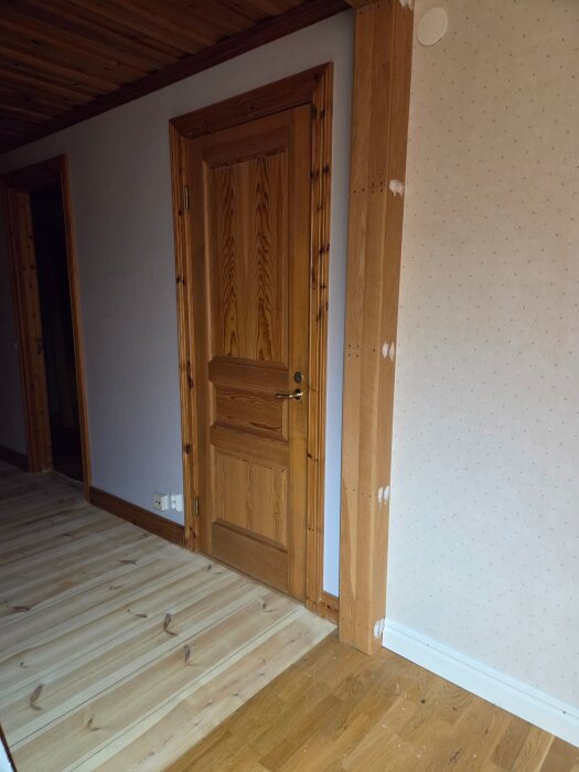 Träpanelvägg med dörr och oslipade spacklade partier i ett hem, potentiellt en äldre innervägg mellan kök och allrum.