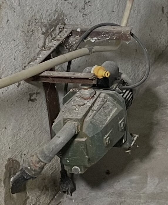 Gammal pump med slang på garagevägg, använd för avloppstryck enligt renoverare, rostig metallstötta och igenpluggad utåtriktad röranslutning.