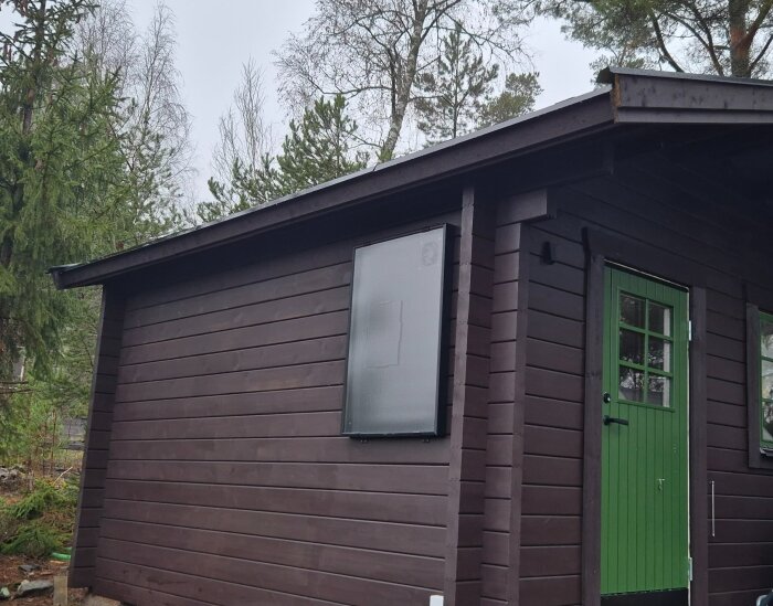 Brun friggebod med en grön dörr och en installerad solventilator på väggen, omgiven av träd.