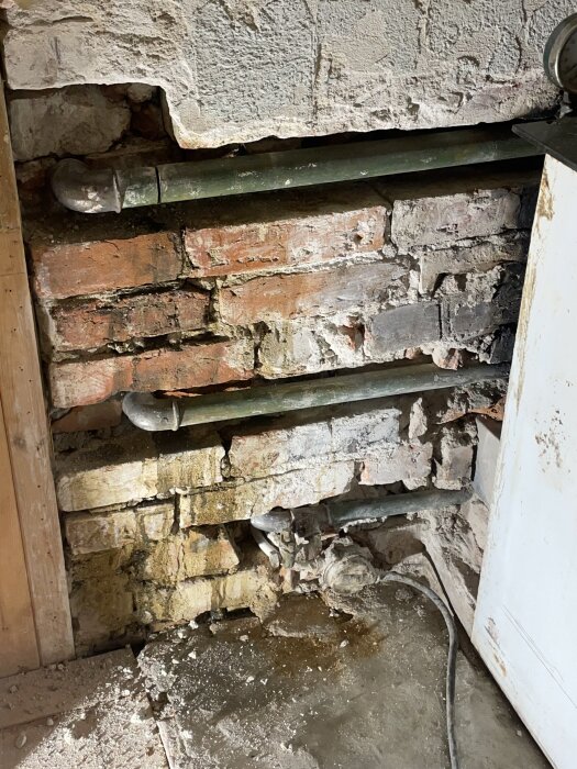 Tre rör på väggen i ett kök, förbundna till en äldre kökspanna, med slitna tegel- och putsomgivningar, grönt rör med vinklade ändar.