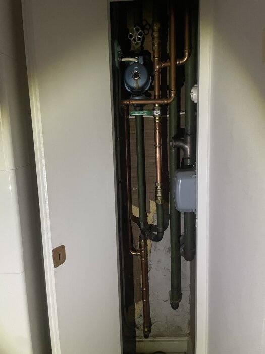 Rördragning med koppar- och gröna rör i ett smalt skåp, inklusive ventil och blå cirkulationspump, del av radiator- och varmvatteninstallation.