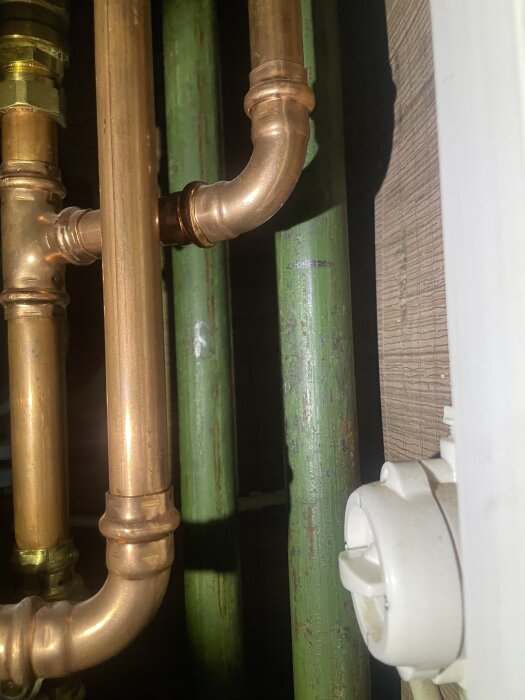 Kopparrör och gröna rör synliga bakom en vägg, del av radiatorkrets, i ett kök med anslutning till äldre Norrahammar kökspanna.